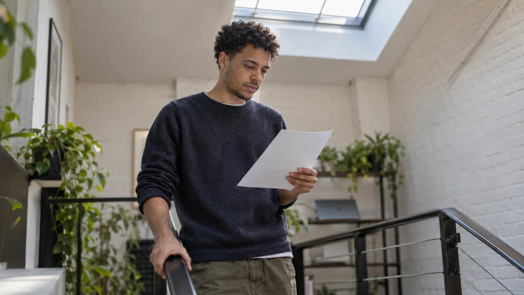 Candid photograph of a mixed-race British man in his twenties with curly hair reading a printed page on a studio stair landing, lit by a skylight.