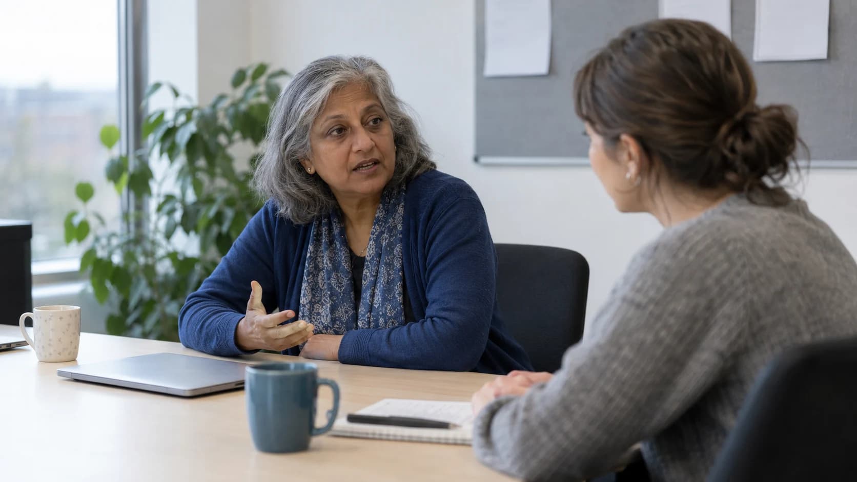 Natural candid photograph of an older South Asian British woman with grey hair explaining something to a colleague across a plain meeting-room table, a closed laptop and mugs between them.