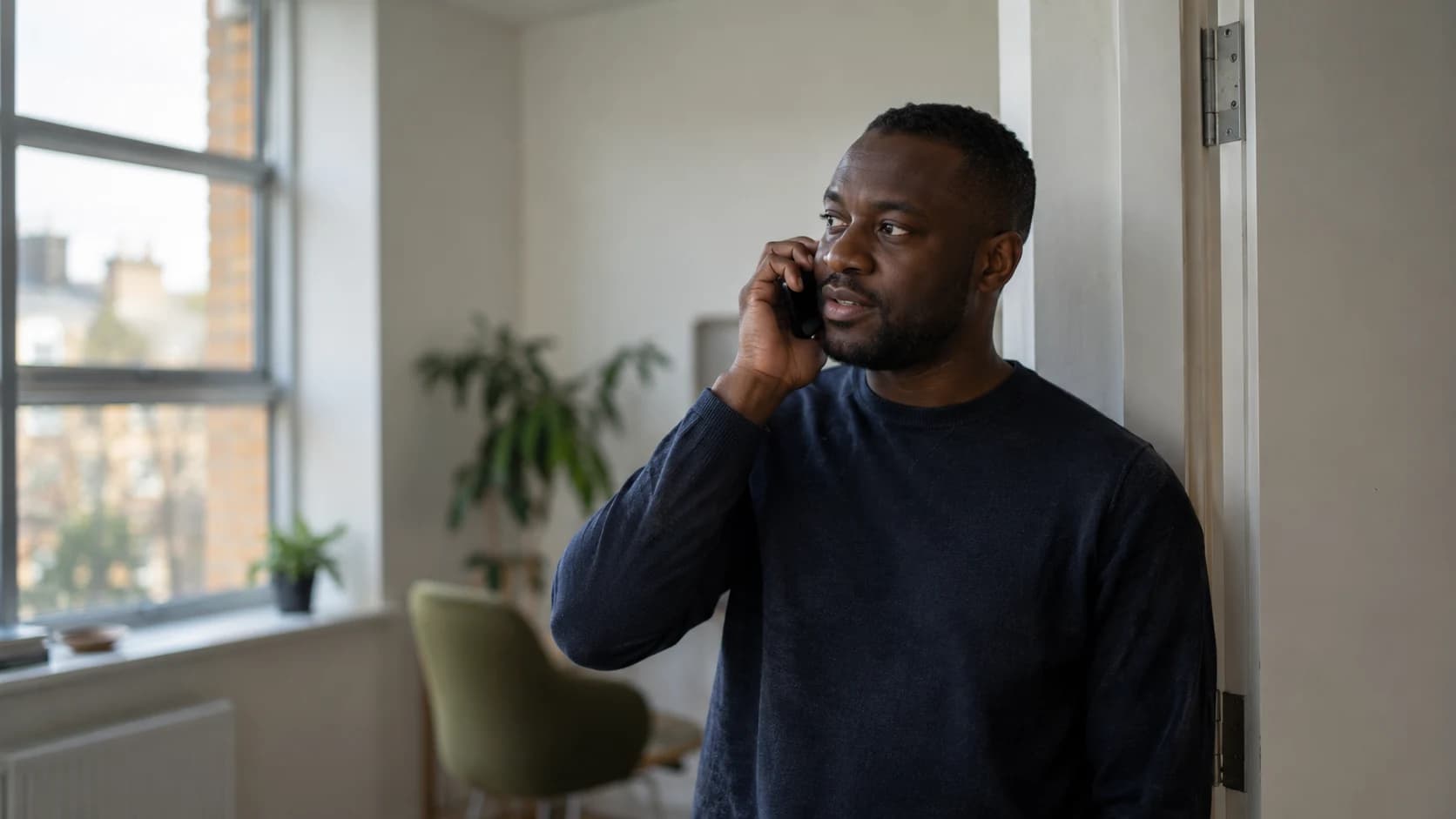 A Black British man in his thirties with close-cropped hair stands in a small UK studio doorway, phone to his ear mid-conversation, in soft daylight.