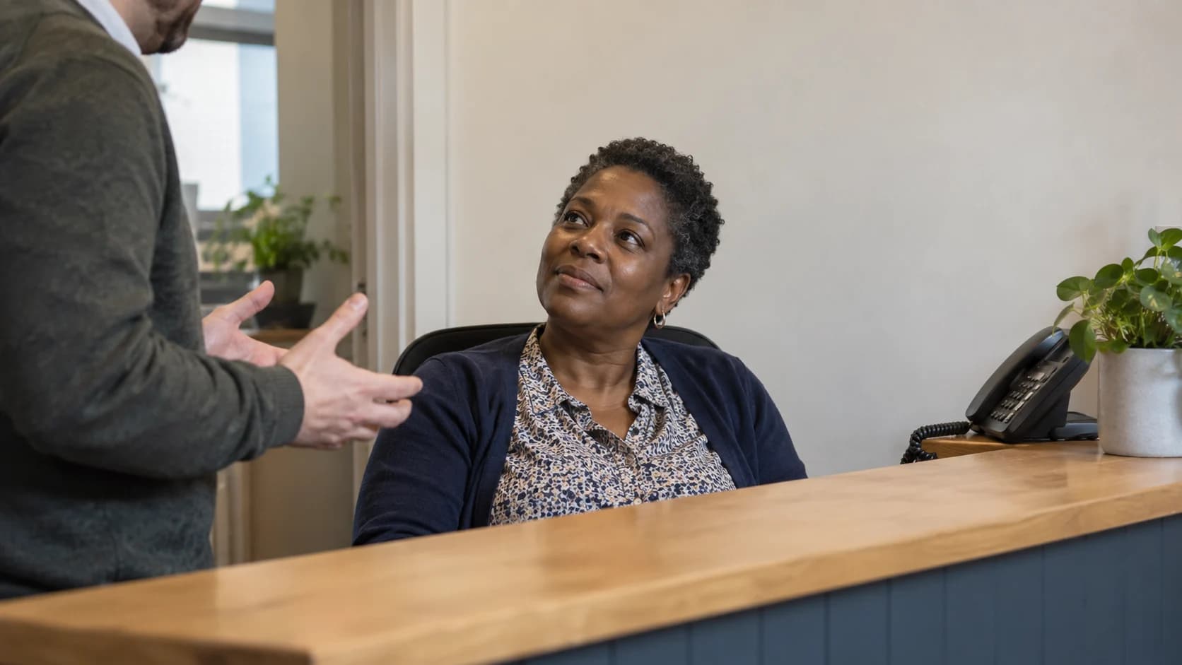 A middle-aged Black British woman with short natural hair at a small business reception desk, leaning back mid-conversation with a colleague explaining a workaround.