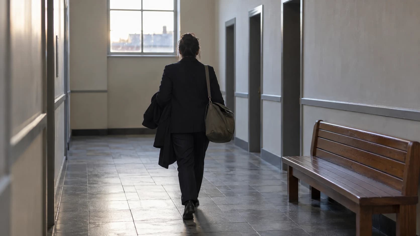 An anonymous professional arriving in a quiet public-building corridor at dawn with a document bag and coat.