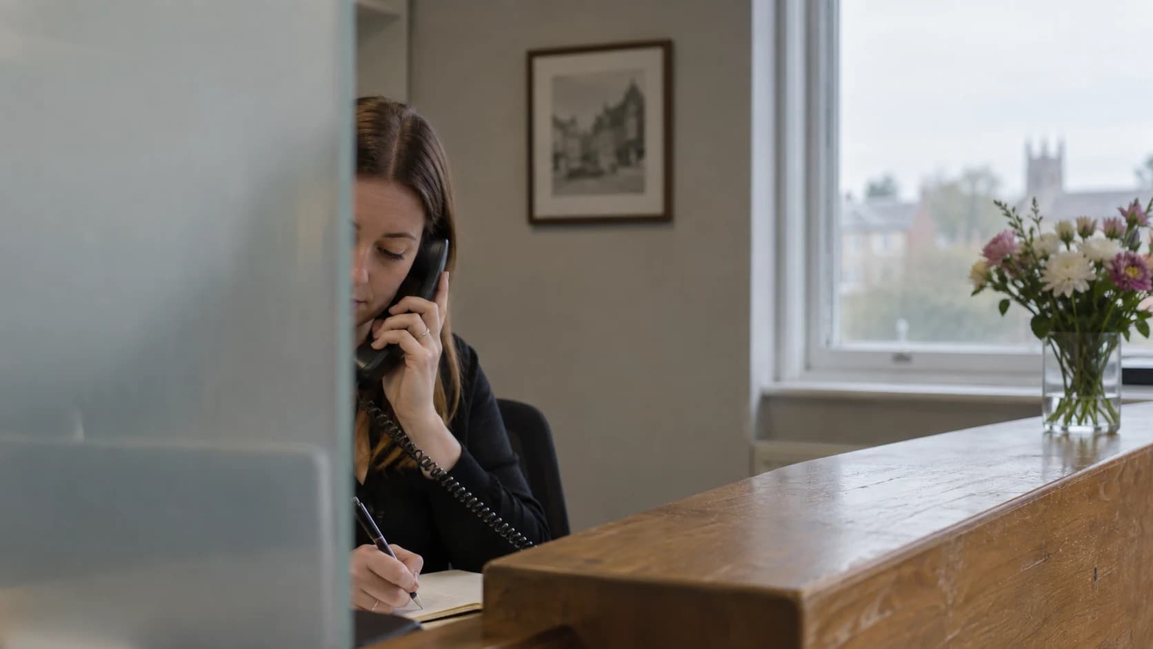 An office receptionist answering a corded phone and taking a note behind a frosted divider.