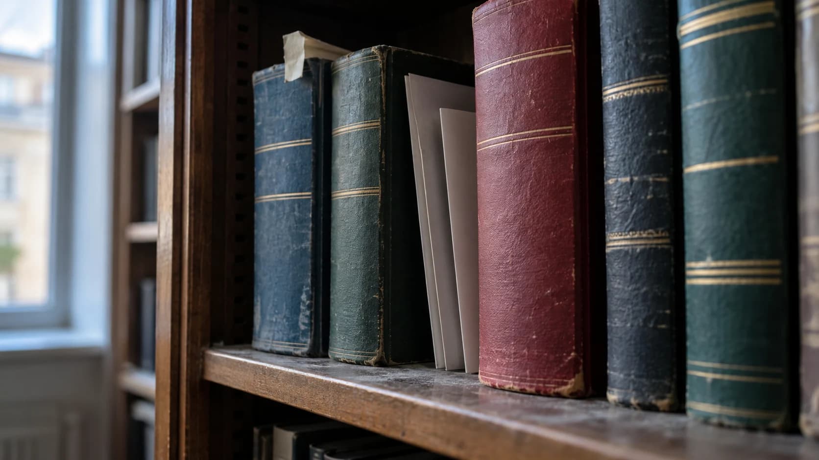 A close angled view of bound legal volumes in several muted colours on a dusty shelf, with plain card leaflets tucked between them, lit by cool daylight.