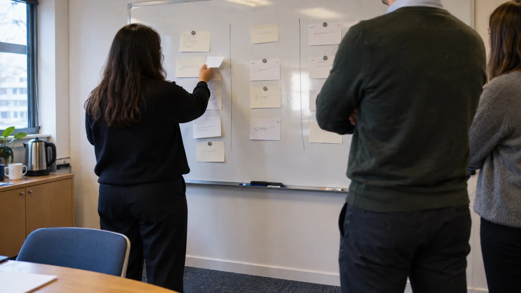 Two or three colleagues seen from behind, arranging scribbled index cards in columns on a whiteboard in a plain meeting room, lit by morning daylight.