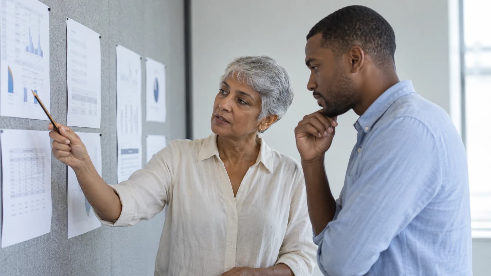 Two colleagues reviewing unreadable printouts pinned to a plain office wall.