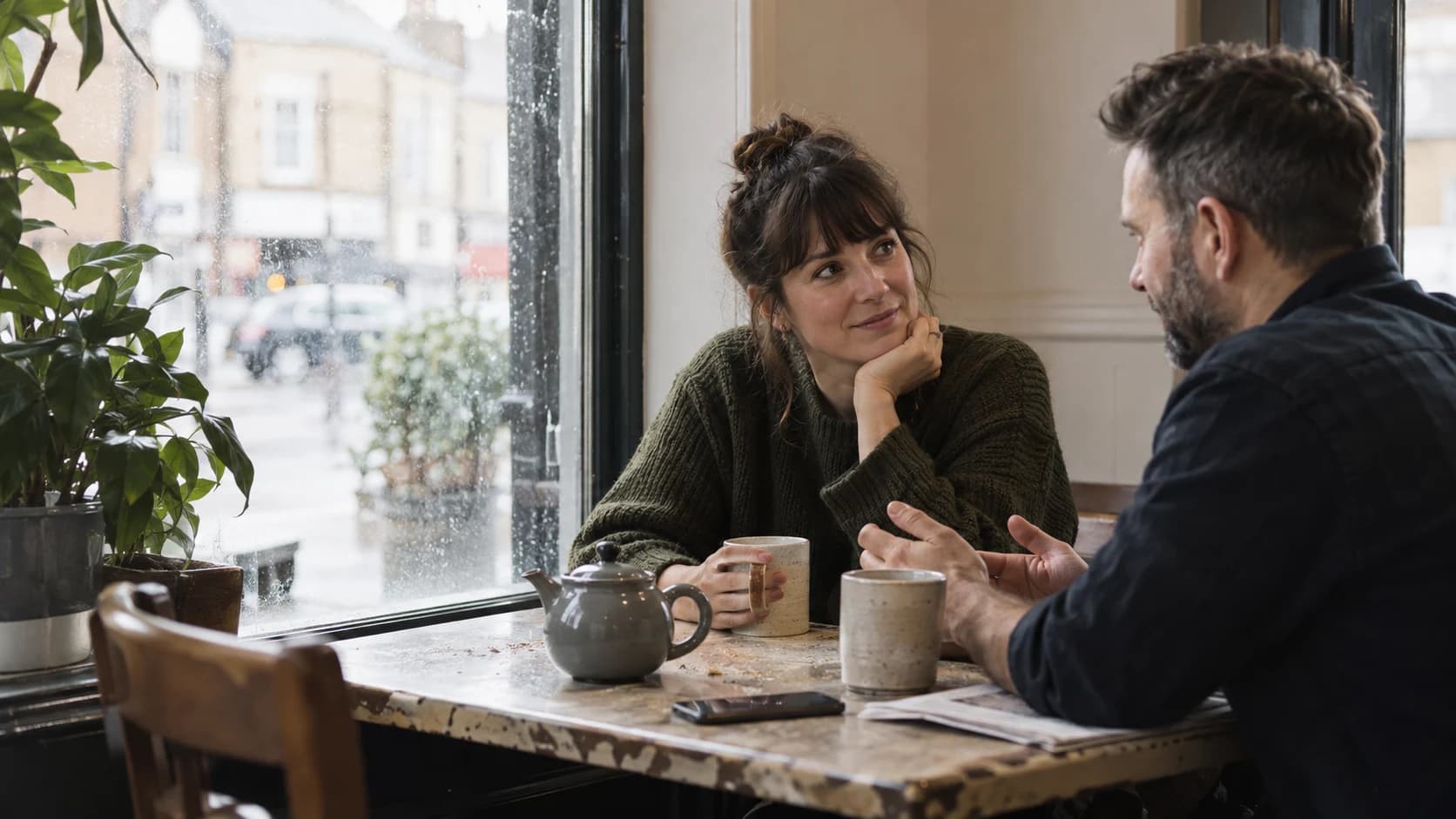 Two people talking over mugs and a face-down phone at a small UK café window table in soft daylight.