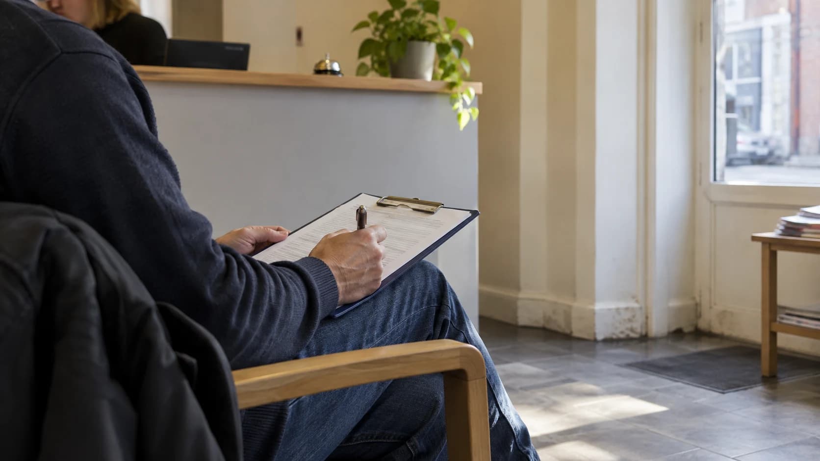 A seated visitor seen from the side, face out of frame, holding a clipboard form in a plain solicitors' reception with a blurred counter behind and daylight on the floor.