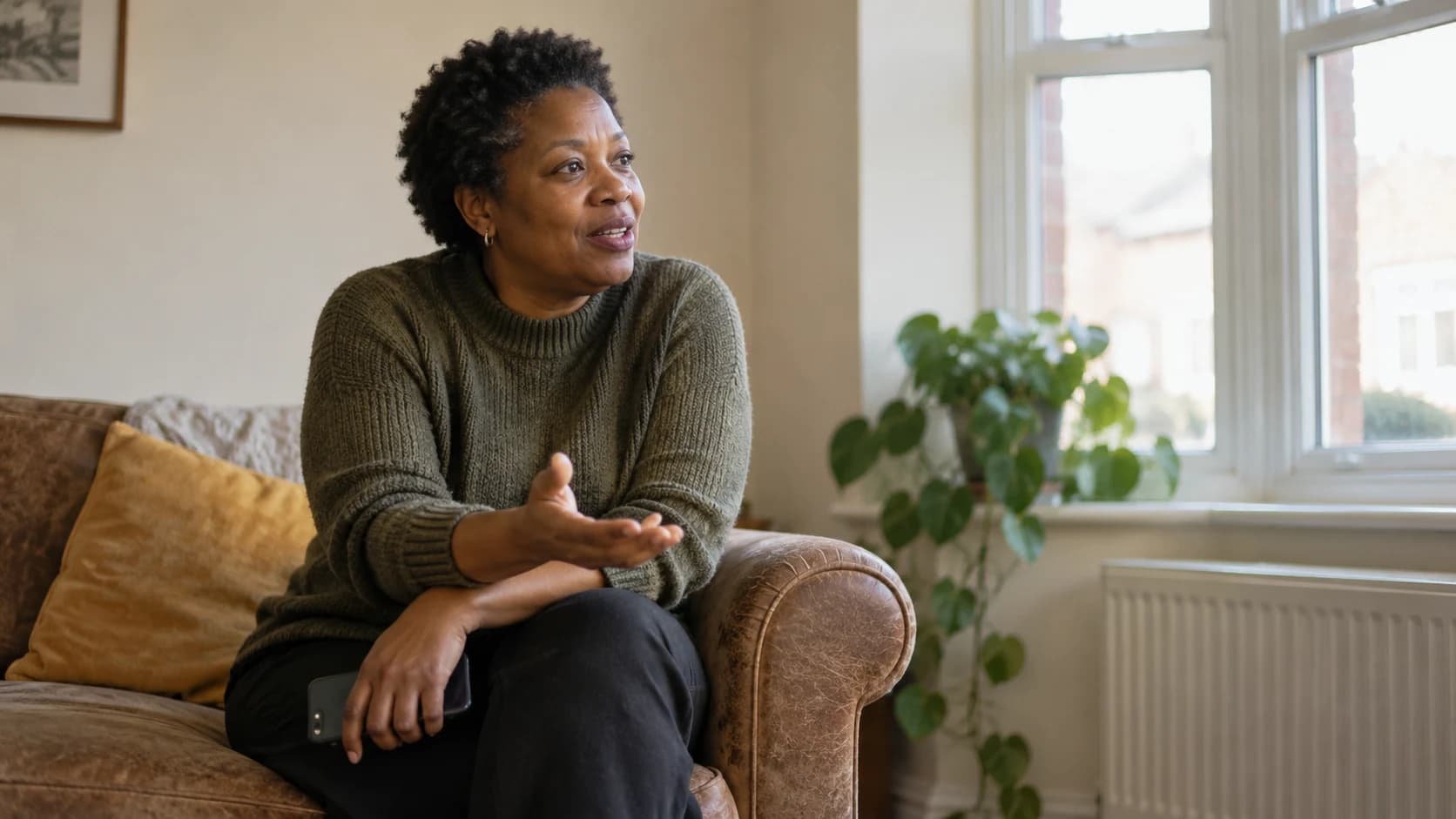 Candid photograph of a middle-aged Black British woman with short natural hair sitting on a sofa arm in a bright UK front room, phone held at her side, talking to someone off-camera.